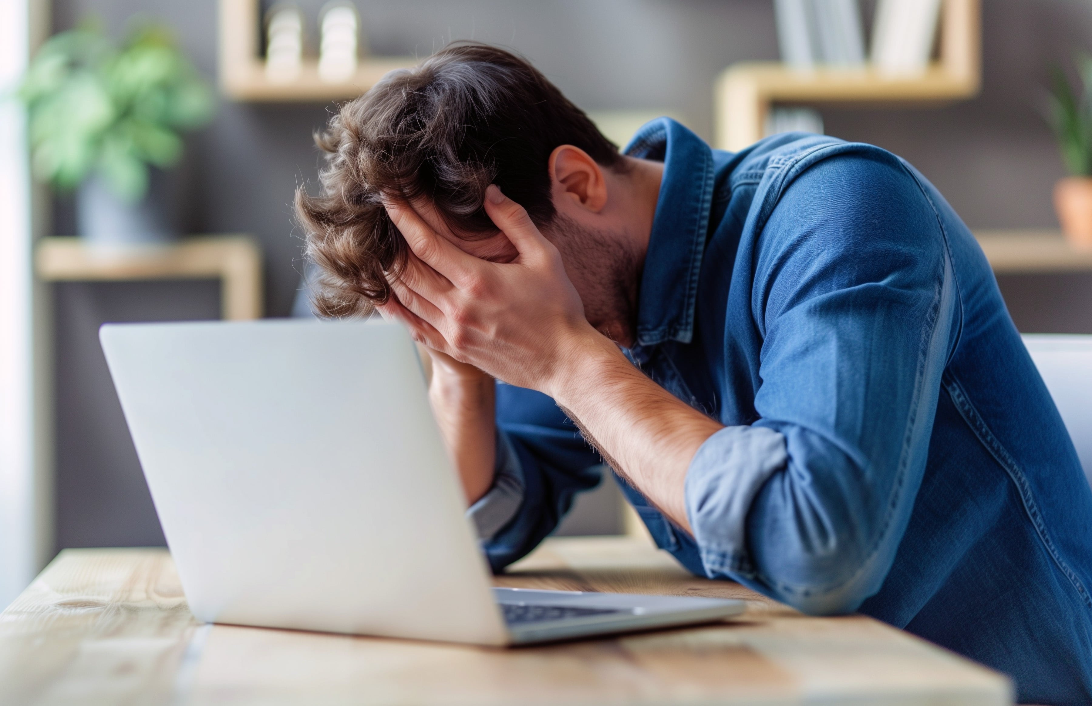 Mann sitzt gestresst vor seinem Laptop und hält die Hände vor sein Gesicht. Erschöpfung.